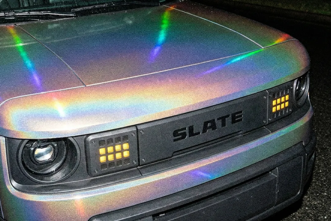 Gros plan de nuit sur le capot et la calandre d’un Slate recouvert d’un film holographique aux reflets arc-en-ciel, phares allumés.